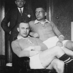 Trójka mężczyzn w mieszkaniu. Dwóch z nich, ubranych w stroje sportowe, siedzi na krześle. Za nimi stoi mężczyzna w ciemnym garniturze.