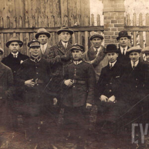 grupa kilkunastu mężczyzn, większość w płaszczach i kapeluszach, dwóch w mundurach wojskowych i rogatywkach, stoi na tle drewnianego płotu rozpiętego między murowanymi z cegły słupkami