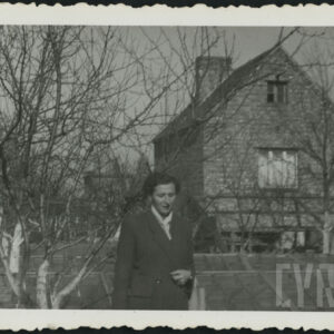 kobieta w płaszczu stoi w ogrodzie wśród drzew owocowych, w głębi murowany, nieotynkowany budynek mieszkalny ze spadzistym dachem i kominem