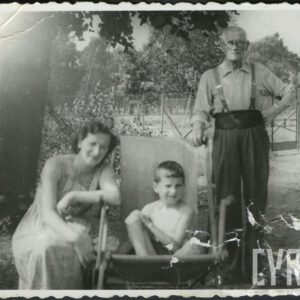 Kobieta siedząca w kucki w ogrodzie, oparta ręką o podłokietnik leżaka, na którym siedzi chłopiec z podkurczonymi nogami, za nimi stoi starszy mężczyzna w okularach trzymający rękę na oparciu leżaka