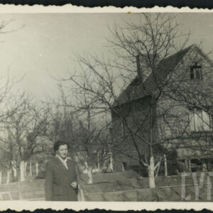 kobieta w płaszczu stoi w ogrodzie, w głębi drzewa owocowe i murowany, nieotynkowany budynek mieszkalny ze spadzistym dachem i kominem