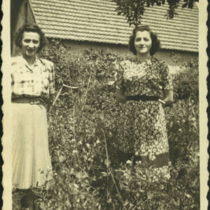 dwie kobiety stoją w ogrodzie, w głębi fragment budynku ze spadzistym dachem pokrytym dachówkami