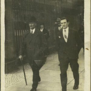 dwóch mężczyzn idących chodnikiem, starszy (z lewej), w kapeluszu i płaszczu, podpiera się laską, młodszy w garniturze, za nimi idą kobieta i mężczyzna, w głębi fragmenty metalowego, wysokiego płotu i budynku z piaskowca