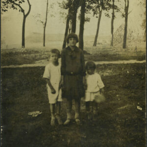 dziewczynka w ciemnej sukience trzyma za ręce dwoje młodszych dzieci w jasnych ubrankach, po lewej chłopca, po prawej dziewczynkę, w głębi kilka drzew