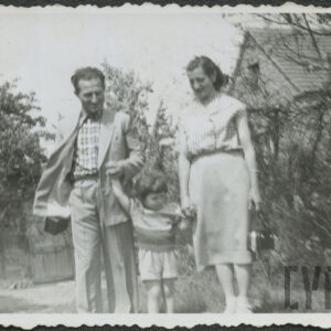 kobieta i mężczyzna stoją w ogrodzie, trzymają pośrodku za ręce małego chłopca, w głębi, z prawej fragment murowanego, nieotynkowanego budynku ze spadzistym dachem