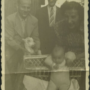 nachylony mężczyzna w jasnym garniturze (z lewej) trzyma za przednie nogi kozę, w głębi starszy mężczyzna w ciemnym garniturze, z prawej młoda kobieta nachylona nad niemowlakiem siedzącym w łóżeczku