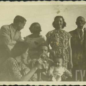 dwie kobiety siedzą, jedna z nich trzyma niemowlę, za nimi dwóch mężczyzn po bokach (z prawej starszy), w środku między mężczyznami dwie kobiety