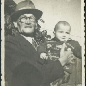 starszy mężczyzna w garniturze, kapeluszu i okularach trzyma na rękach małe dziecko