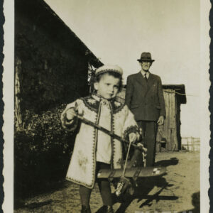 mały chłopiec w haftowanym żakiecie i czapce na głowie, w rękach trzyma model samolotu, za nim starszy mężczyzna w garniturze i kapeluszu, za nimi z lewej ściana murowanego budynku, w głębi fragment budynku drewnianego