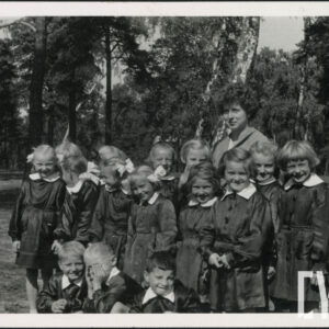 kilkanaścioro dzieci w ciemnych fartuszkach z białymi kołnierzykami, z czego trzech chłopców kuca, za nimi kobieta, wszyscy stoją na tle drzew