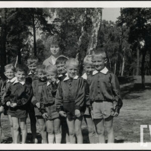 dziesięciu chłopców w ciemnych fartuszkach z białymi kołnierzykami, za nimi kobieta, wszyscy stoją na tle drzew