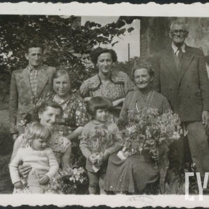 dwie kobiety siedzą, jedna z nich (z prawej) trzyma na kolanach kwiaty, przed kobietą z lewej siedzi na trawie kobieta i trzyma dziecko, pomiędzy nimi stoi drugie dziecko, za nimi stoją po bokach dwaj mężczyźni, pomiędzy nimi kobieta, w głębi gałąź drzewa, z prawej fragment  ściany murowanego budynku z oknem