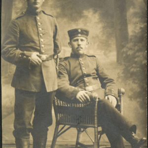 dwóch mężczyzn w mundurach, jeden stoi (z lewej), drugi siedzi na krześle, w atelier na tle tapety z krajobrazem z drzewami