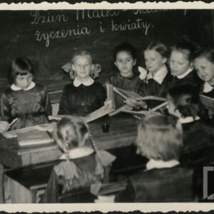 nauczycielka i uczennice w mundurkach z białymi kołnierzykami siedzą wokół szkolnej ławki, za ich plecami wisi szkolna tablica