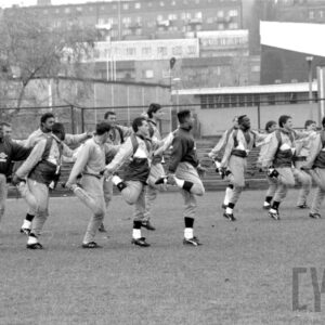 grupa młodych mężczyzn w strojach sportowych wykonująca ćwiczenia fizyczne na boisku do gry w piłkę nożną na tle ławek, ogrodzenia z siatki i budynków mieszkalnych