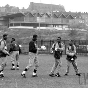 grupa młodych mężczyzn w strojach sportowych wykonująca ćwiczenia fizyczne na boisku do gry w piłkę nożną na tle ławek i budynków