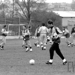 grupa młodych mężczyzn w strojach sportowych wykonująca ćwiczenia fizyczne na boisku do gry w piłkę nożną na tle ławek, ogrodzenia z siatki i budynków mieszkalnych