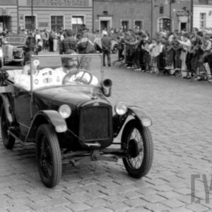 mężczyźni w strojach z dawnej epoki jadący w zabytkowym samochodzie po bruku; w tle grupa ludzi obserwująca paradę oraz zabytkowe budynki