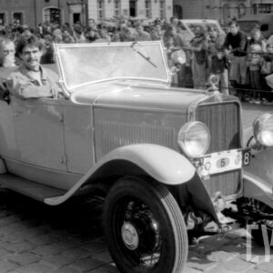 mężczyźni jadący w zabytkowym samochodzie po bruku; w tle grupa ludzi obserwująca paradę oraz zabytkowe budynki