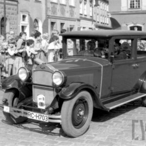 mężczyźni w strojach z dawnej epoki jadący w zabytkowym samochodzie po bruku; w tle grupa ludzi obserwująca paradę oraz zabytkowe budynki