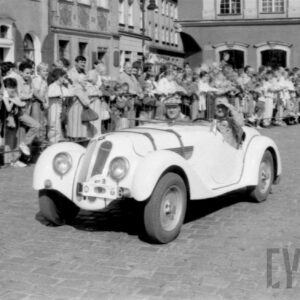 mężczyźni w strojach z dawnej epoki jadący w zabytkowym samochodzie po bruku; w tle grupa ludzi obserwująca paradę oraz zabytkowe budynki