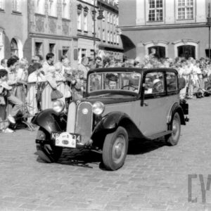 grupa ludzi w strojach z dawnej epoki jadąca w zabytkowym samochodzie po bruku; w tle grupa ludzi obserwująca paradę oraz zabytkowe budynki