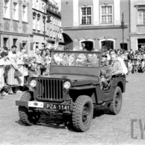 grupa ludzi w strojach z dawnej epoki jadąca w zabytkowym samochodzie po bruku; w tle grupa ludzi obserwująca paradę oraz zabytkowe budynki