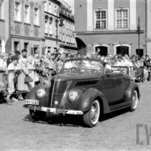 grupa ludzi w strojach z dawnej epoki jadąca w zabytkowym samochodzie po bruku; w tle grupa ludzi obserwująca paradę oraz zabytkowe budynki
