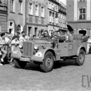 grupa ludzi w strojach z dawnej epoki jadąca w zabytkowym samochodzie po bruku; w tle grupa ludzi obserwująca paradę oraz zabytkowe budynki