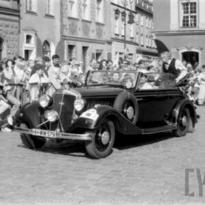 grupa ludzi w strojach z dawnej epoki jadąca w zabytkowym samochodzie po bruku; w tle grupa ludzi obserwująca paradę oraz zabytkowe budynki