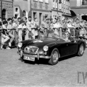 grupa ludzi w strojach z dawnej epoki jadąca w zabytkowym samochodzie po bruku; w tle grupa ludzi obserwująca paradę oraz zabytkowe budynki