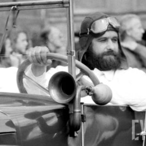 mężczyzna w średnim wieku noszący brodę w czapce pilotce i goglach z dawnej epoki jadący zabytkowym samochodem