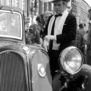 starszy mężczyzna w stroju z dawnej epoki stojący przy zabytkowym samochodzie na tle kamienic i grupy ludzi przyglądających się paradzie