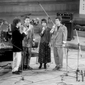 trzech mężczyzn i jedna kobieta śpiewający do trzymanych w dłoniach mikrofonów; po bokach dwóch kamerzystów; w tle sprzęt muzyczny