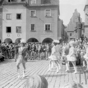 grupa kobiet prezentujących układ taneczny w tle grupa gapiów oraz zespół orkiestry dętej; na drugim planie figura na pręgierzu oraz kamienice