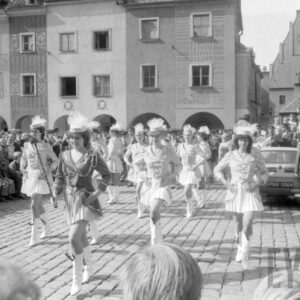 grupa kobiet prezentujących układ taneczny w tle grupa gapiów oraz zespół orkiestry dętej; na drugim planie figura na pręgierzu oraz kamienice