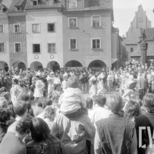 grupa kobiet prezentujących układ taneczny w tle grupa gapiów oraz zespół orkiestry dętej; na drugim planie figura na pręgierzu oraz kamienice