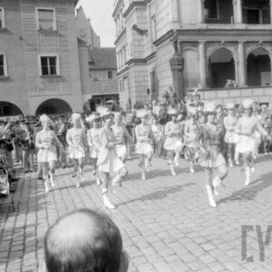 grupa kobiet prezentujących układ taneczny w tle grupa gapiów oraz zespół orkiestry dętej; na drugim planie figura na pręgierzu oraz kamienice