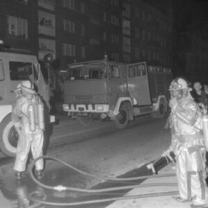 dwóch strażaków w strojach gaśniczych w czasie akcji gaśniczej przy wozach strażackich na tle bloków, obok stoi grupa gapiów