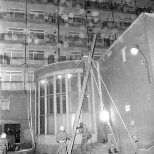 trzech strażaków z drabiną przystawioną do budynku, na drabinę wspina się kolejny ze strażaków, na drugim planie blok mieszkalny na którego balkonach stoją przyglądający się akcji strażaków gapie