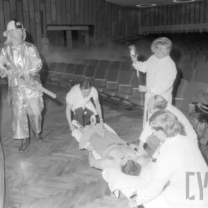 strażak i grupa medyków udzielająca pomocy poszkodowanemu mężczyźnie leżącemu na noszach w sali kinowej, na tle rzędów foteli kinowych