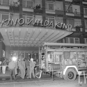 wóz strażacki zaparkowany na ulicy naprzeciwko wejścia do budynku, przy wozie stoją strażacy i grupa przypadkowych ludzi