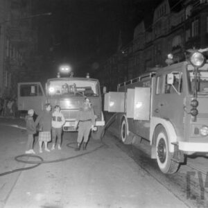 strażak i grupa gapiów przy dwóch wozach strażackich stojących na ulicy między kamienicami