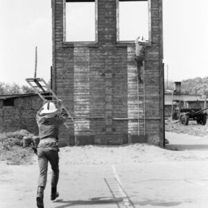 dwóch mężczyzn w kombinezonach i hełmach strażackich; jeden z nich biegnie trzymając w rękach drabinę, drugi wspina się po drabinie na drewnianą ścianę do ćwiczeń