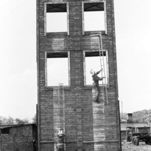 dwóch mężczyzn w kombinezonach i hełmach strażackich; jeden z nich biegnie trzymając w rękach drabinę, drugi wspina się po drabinie na drewnianą ścianę do ćwiczeń