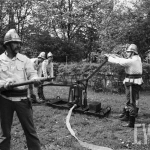 grupa mężczyzn w dawnych strojach strażackich na tle drzew; jeden z nich trzyma węża gaśniczego, z tyłu czterech kolejnych pompuje wodę przy pomocy historycznej pompy
