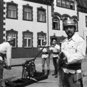 grupa mężczyzn w dawnych strojach strażackich; jeden z nich trzyma węża gaśniczego, z tyłu czterech kolejnych pompuje wodę przy pomocy historycznej pompy; w tle budynek ceglany