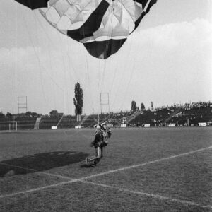 młody mężczyzna w kombinezonie z paralotnią na stadionie, w tle trybuny i drzewa