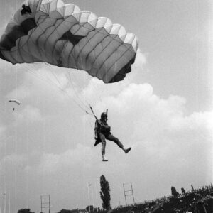 młody mężczyzna w kombinezonie z paralotnią lądujący stadionie, w tle trybuny i drzewa