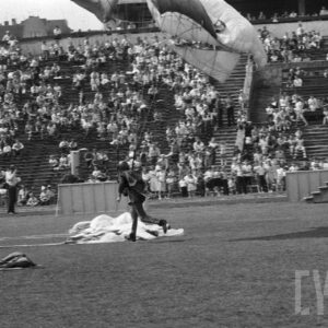 młody mężczyzna w kombinezonie z paralotnią na stadionie, w towarzystwie trójki mężczyzn, w tle trybuny z grupą ludzi obserwujących pokaz